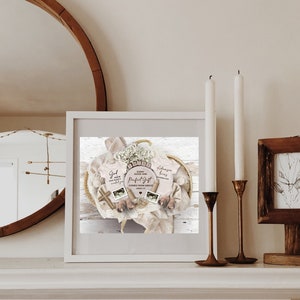 May include: A framed print of a baby shower gift basket with a white background. The basket contains two pink baby onesies, one with the text "God made you so wonderfully" and the other with the text "Ephesians 1:4", a small wooden cross, a white blanket, and a white flower arrangement. The text "Every good and perfect gift comes from above" is written on a wooden sign.