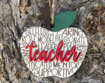 Può includere: Un ornamento di legno bianco a forma di mela con la parola "teacher" in lettere rosse circondata da parole che descrivono le qualità di un insegnante, come "caring", "passionate", "leader", "mentor" e "supportive".
