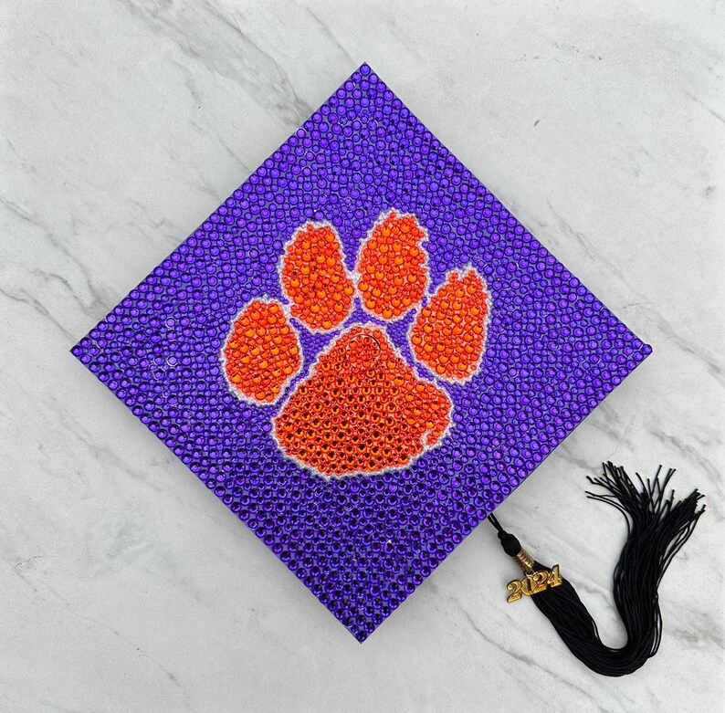 May include: Purple graduation cap decorated with orange and white rhinestones in the shape of a paw print. The cap has a black tassel with a gold charm that says "2024".