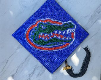 May include: A blue graduation cap decorated with rhinestones featuring the University of Florida Gators mascot, an alligator with an orange and white outline. The cap has a black tassel with a gold 2024 charm.