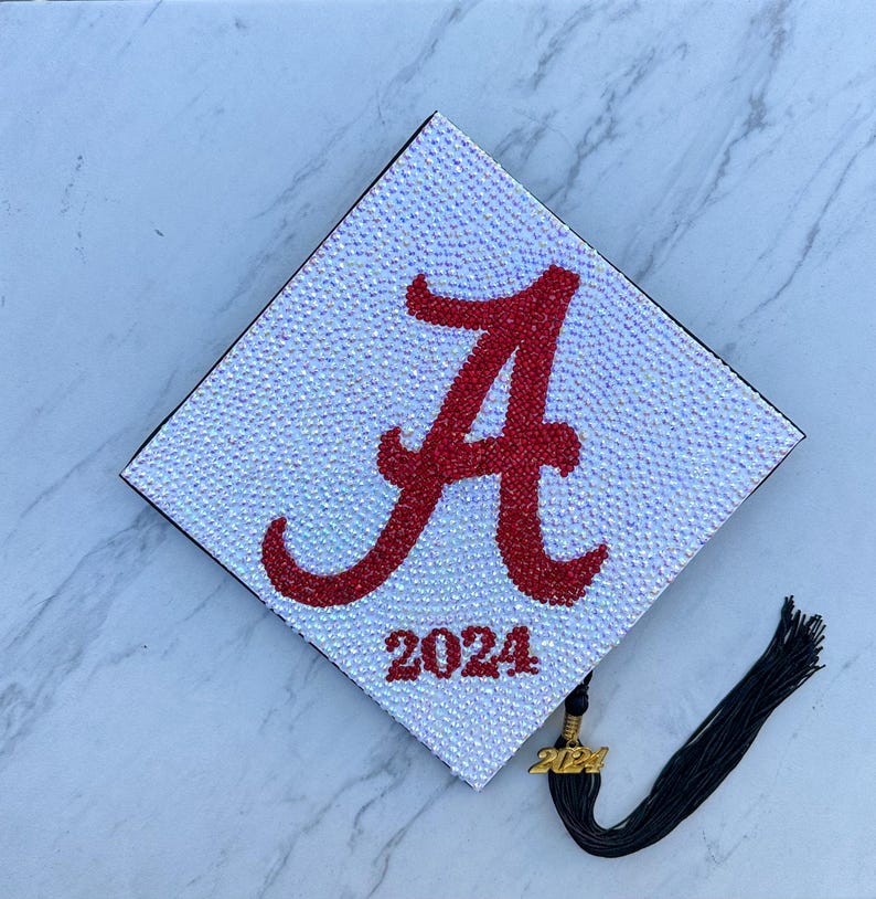 May include: A white graduation cap covered in iridescent rhinestones with a red "A" and the year "2024" in rhinestones. A black tassel with a gold 2024 charm is attached.