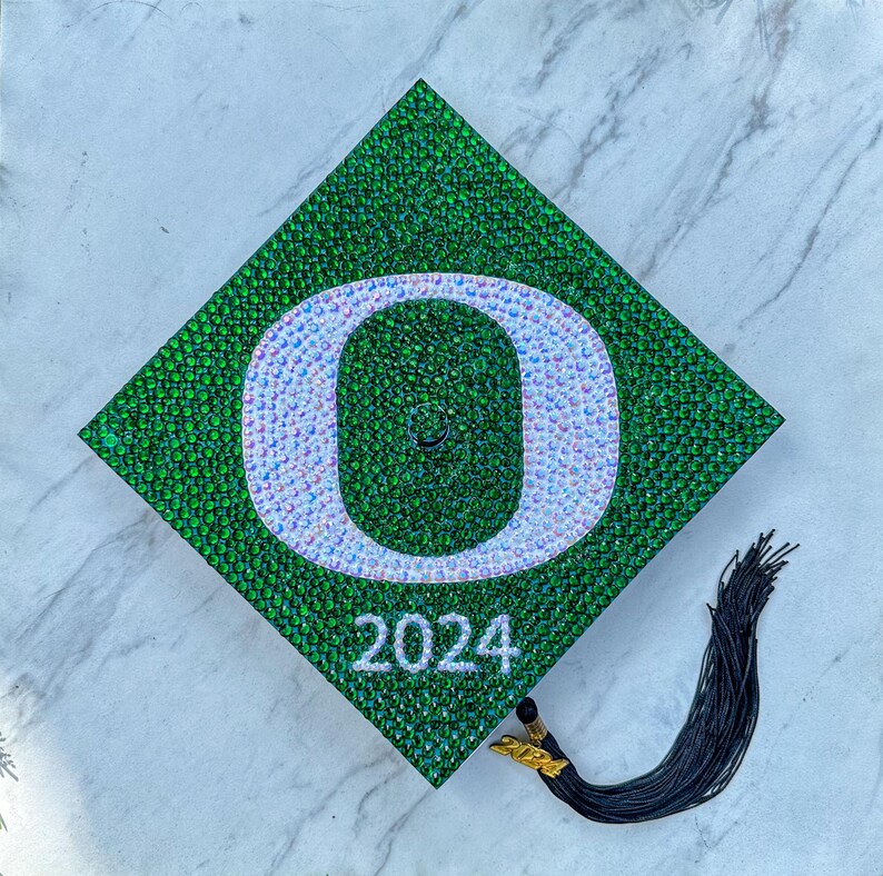 May include: Green graduation cap decorated with rhinestones in the shape of a white letter "O" with a white background. The year 2024 is written in white below the letter. A black tassel hangs from the cap.
