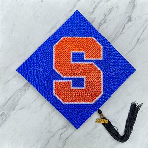 May include: A blue and orange graduation cap with a large letter 'S' made of rhinestones. The cap has a gold tassel with the year '2021' on it.
