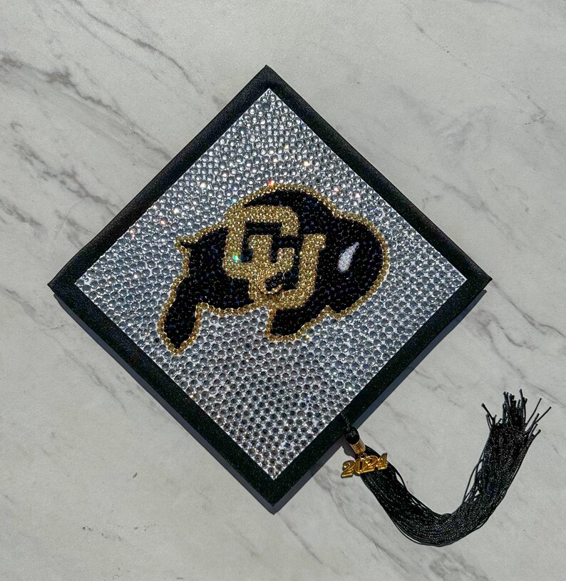 May include: A black graduation cap covered in rhinestones with a gold and black University of Wyoming logo. The cap has a black tassel with a gold 2021 charm.