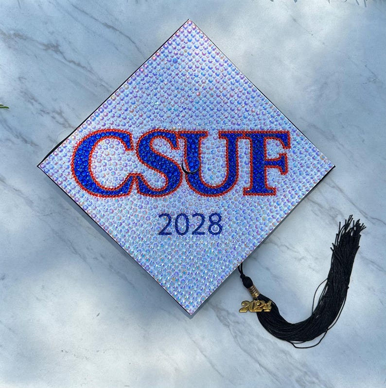 May include: A white graduation cap decorated with blue and red rhinestones spelling "CSUF" and the year "2028". A black tassel hangs from the cap.