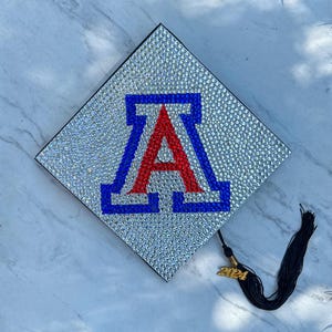 May include: A graduation cap covered in clear rhinestones with a blue and red "A" in the center. The tassel is black with a gold 2024 charm.