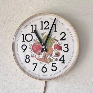 Könnte beinhalten: Runde Wanduhr mit einem hellen Holzrahmen. Das Zifferblatt zeigt ein Stillleben aus Gemüse und Obst, darunter Tomaten, Artischocken und Pilze. Schwarze Ziffern markieren die Stunden. Die Zeiger der Uhr sind schwarz. Der Markenname "Westclox" ist auf dem Zifferblatt aufgedruckt.