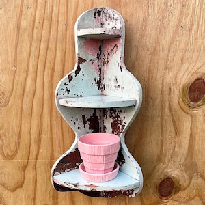May include: A distressed, vintage-style corner shelf with three tiers, painted in faded blue, pink, and white. A pink ceramic flower pot and saucer sit on the bottom shelf. The shelf is mounted on a wooden wall.