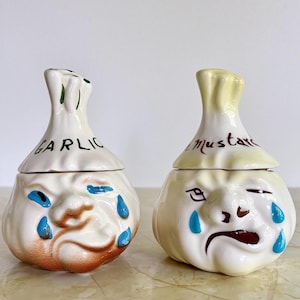 Puede incluir: Dos tarros de cerámica para ajo y mostaza con caras. El tarro de ajo es blanco con detalles marrones y la palabra "GARLIC" en verde. El tarro de mostaza es blanco con detalles amarillos y la palabra "Mustard" en marrón. Ambos tienen lágrimas azules.