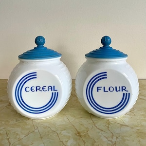 Puede incluir: Dos botes de cerámica blanca con tapas azules. Uno dice "CEREAL" y el otro "FLOUR" en azul. Los botes tienen un diseño acanalado, ideales para el almacenamiento en la cocina.