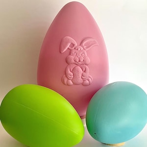 May include: A pink, egg-shaped Easter decoration with a carved bunny holding an egg, flanked by a green and a blue egg. The bunny is holding an egg. The eggs are in shades of green and blue.