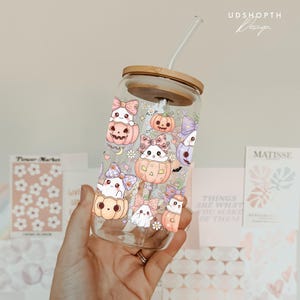 May include: A clear glass tumbler with a bamboo lid and straw, decorated with a Halloween-themed design. The design features cute ghost characters in pumpkins, adorned with bows. The text "UDSHOPTH Design" is visible on the upper right.