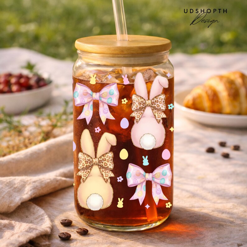 May include: A clear glass tumbler with a wooden lid and straw, filled with a dark liquid. The glass is decorated with Easter-themed bunny designs and bows. The background includes a picnic setting with a croissant and coffee beans.