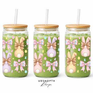 May include: Three glass tumblers filled with a green liquid, each topped with a wooden lid and a straw. The tumblers are decorated with Easter bunny designs, including bows and bunny tails. The design includes pastel colors and a leopard print.