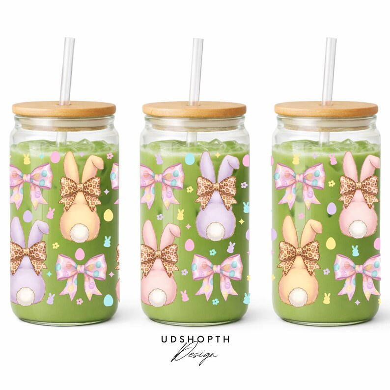May include: Three glass tumblers filled with a green liquid, each topped with a wooden lid and a straw. The tumblers are decorated with Easter bunny designs, including bows and bunny tails. The design includes pastel colors and a leopard print.