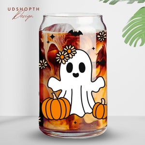 May include: A clear glass can with a white ghost wearing a flower crown and holding a pumpkin. The can is filled with a brown liquid and has a white and black bat design on the side.