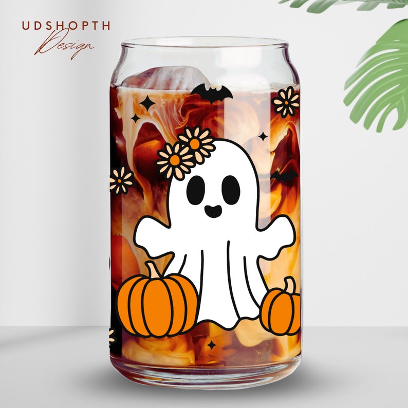 May include: A clear glass can with a white ghost wearing a flower crown and holding a pumpkin. The can is filled with a brown liquid and has a white and black bat design on the side.