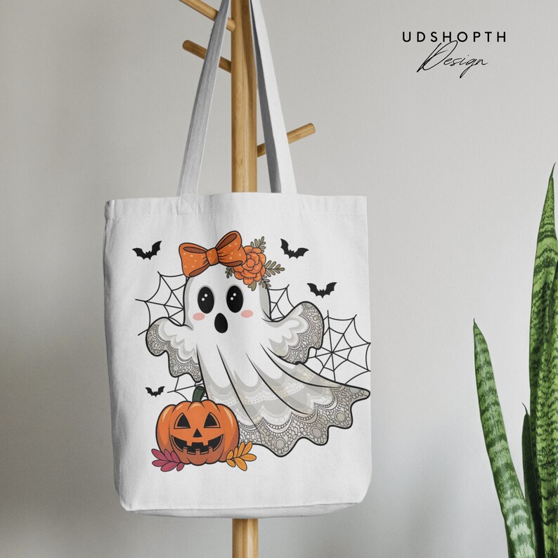 Pode incluir: Sacola de tecido branca com um fantasma de desenho animado fofo usando um la&ccedil;o e uma faixa de flores. O fantasma est&aacute; cercado por teias de aranha e morcegos. H&aacute; uma ab&oacute;bora esculpida com a forma de uma caveira no canto inferior esquerdo.