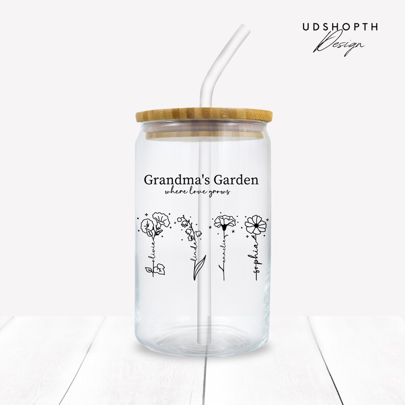 K&ouml;nnte beinhalten: Ein klares Glasgef&auml;&szlig; mit Bambusdeckel und Strohhalm. Das Gef&auml;&szlig; hat ein schwarzes Design mit der Aufschrift "Grandma's Garden where love grows" und zeigt vier stilisierte Blumen mit darauf geschriebenen Namen: "Blessing", "Linda", "Kamelia" und "Sophia".