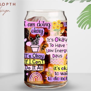 Peut inclure: Un verre transparent en forme de canette avec divers autocollants de texte aux couleurs pastel. Les autocollants comportent des phrases telles que "I am doing okay" et "It's okay to have low energy days". La tasse pr&eacute;sente &eacute;galement une petite plante en pot, un arc-en-ciel et des tournesols.