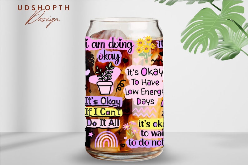 Peut inclure: Un verre transparent en forme de canette avec divers autocollants de texte aux couleurs pastel. Les autocollants comportent des phrases telles que "I am doing okay" et "It's okay to have low energy days". La tasse pr&eacute;sente &eacute;galement une petite plante en pot, un arc-en-ciel et des tournesols.