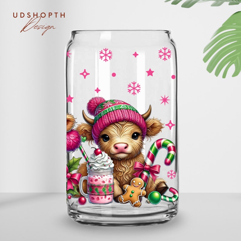 K&ouml;nnte beinhalten: Ein klares Glasgef&auml;&szlig; mit einem rosa und gr&uuml;nen Design, das eine schottische Hochlandkuh zeigt, die einen rosa und gr&uuml;nen Hut tr&auml;gt, einen Zuckerstock und einen Milchshake h&auml;lt. Das Design enth&auml;lt auch einen Lebkuchenmann und Schneeflocken.