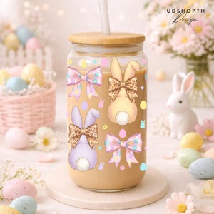 May include: A clear glass tumbler with a wooden lid and a straw, decorated with Easter-themed bunny designs and bows. The tumbler contains a light brown liquid. The design includes pastel colors and the text "UDSHOPTH Design".