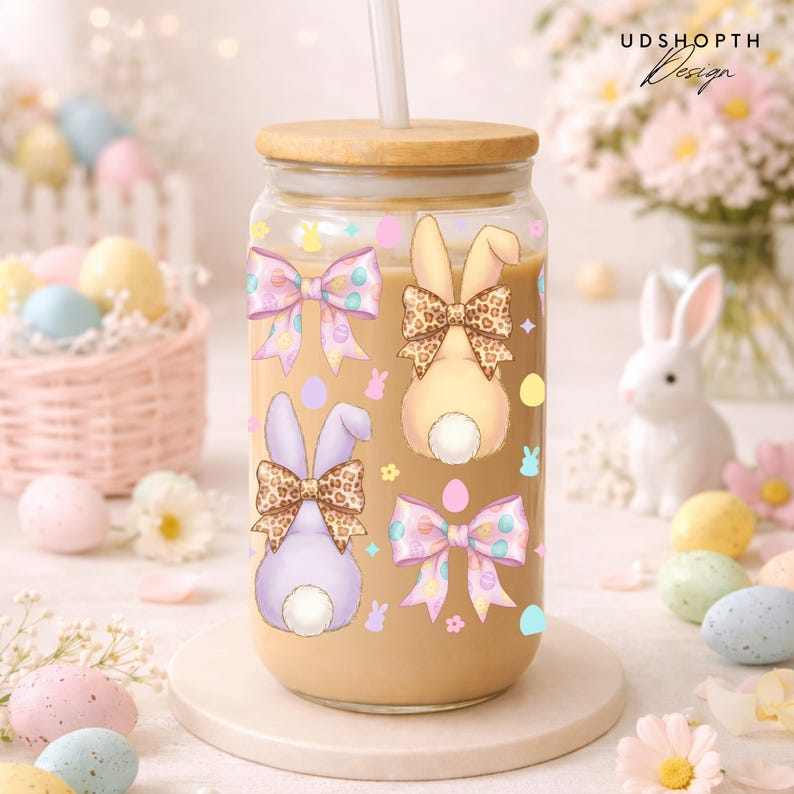 May include: A clear glass tumbler with a wooden lid and a straw, decorated with Easter-themed bunny designs and bows. The tumbler contains a light brown liquid. The design includes pastel colors and the text "UDSHOPTH Design".