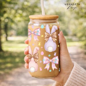 May include: A clear glass tumbler with a bamboo lid and straw, featuring Easter-themed illustrations. The design includes pastel-colored bows, bunny silhouettes, and eggs. The tumbler is filled with a light brown liquid.