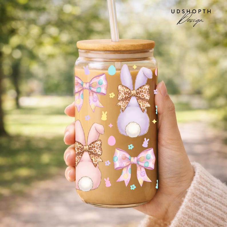 May include: A clear glass tumbler with a bamboo lid and straw, featuring Easter-themed illustrations. The design includes pastel-colored bows, bunny silhouettes, and eggs. The tumbler is filled with a light brown liquid.