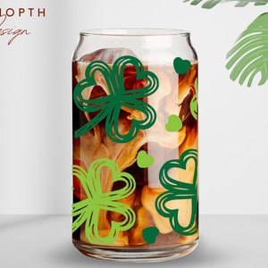 May include: A clear glass can with a brown liquid and green shamrock designs. The can is filled with a brown liquid and has a white foam on top. The shamrocks are green and have a white outline.
