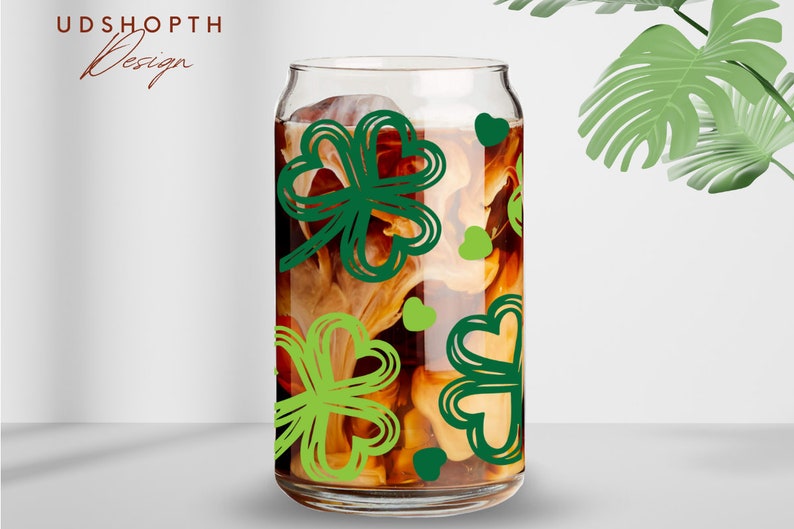 May include: A clear glass can with a brown liquid and green shamrock designs. The can is filled with a brown liquid and has a white foam on top. The shamrocks are green and have a white outline.