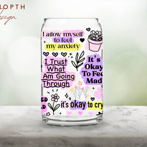 Peut inclure: Un verre transparent en forme de canette avec divers graphiques et textes color&eacute;s. Le texte comprend des phrases telles que "Je m'autorise &agrave; ressentir mon anxi&eacute;t&eacute;" et "C'est ok de pleurer". Le design pr&eacute;sente des c&oelig;urs, des fleurs et une plante succulente en pot.