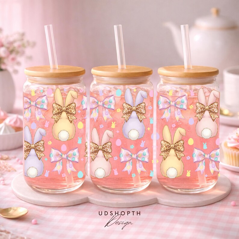 May include: Three glass tumblers with wooden lids and straws, filled with pink liquid. Each tumbler features a bunny design with a leopard print bow, surrounded by bows and Easter eggs. The text "UDSHOPTH Design" is at the bottom.