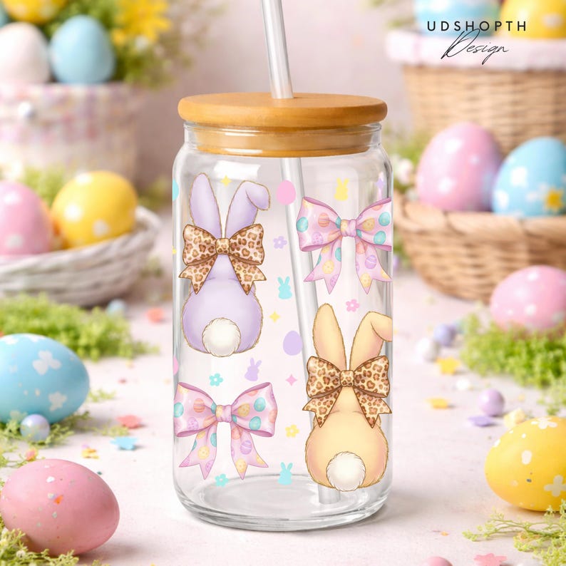 May include: A clear glass tumbler with a bamboo lid and straw, decorated with pastel Easter bunny illustrations and bows. The bunnies have leopard print bows. The text "UDSHOPTH Design" is visible on the top right.