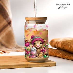 K&ouml;nnte beinhalten: Ein Glasgef&auml;&szlig; mit einem Holzdeckel und einem festlichen Design mit zwei cartoonartigen schottischen Hochlandrindern, die rosa H&uuml;te tragen und Zuckerstangen halten. Das Gef&auml;&szlig; ist mit einer braunen Fl&uuml;ssigkeit und Eisw&uuml;rfeln gef&uuml;llt. Das Design beinhaltet auch einen Lebkuchenmann, ein Schneeflockenmuster und den Text "UDSHOPTH Design".