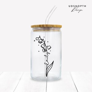 K&ouml;nnte beinhalten: Ein durchsichtiger Glasbecher mit einem Bambusdeckel und einem durchsichtigen Plastikstrohhalm. Der Becher ist mit einer schwarzen Linienzeichnung einer Maigl&ouml;ckchenblume verziert, auf deren Stiel der Name "Emily" steht. Der Becher steht auf einer wei&szlig;en Holzoberfl&auml;che.