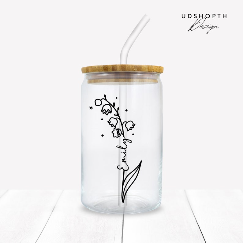 K&ouml;nnte beinhalten: Ein durchsichtiger Glasbecher mit einem Bambusdeckel und einem durchsichtigen Plastikstrohhalm. Der Becher ist mit einer schwarzen Linienzeichnung einer Maigl&ouml;ckchenblume verziert, auf deren Stiel der Name "Emily" steht. Der Becher steht auf einer wei&szlig;en Holzoberfl&auml;che.