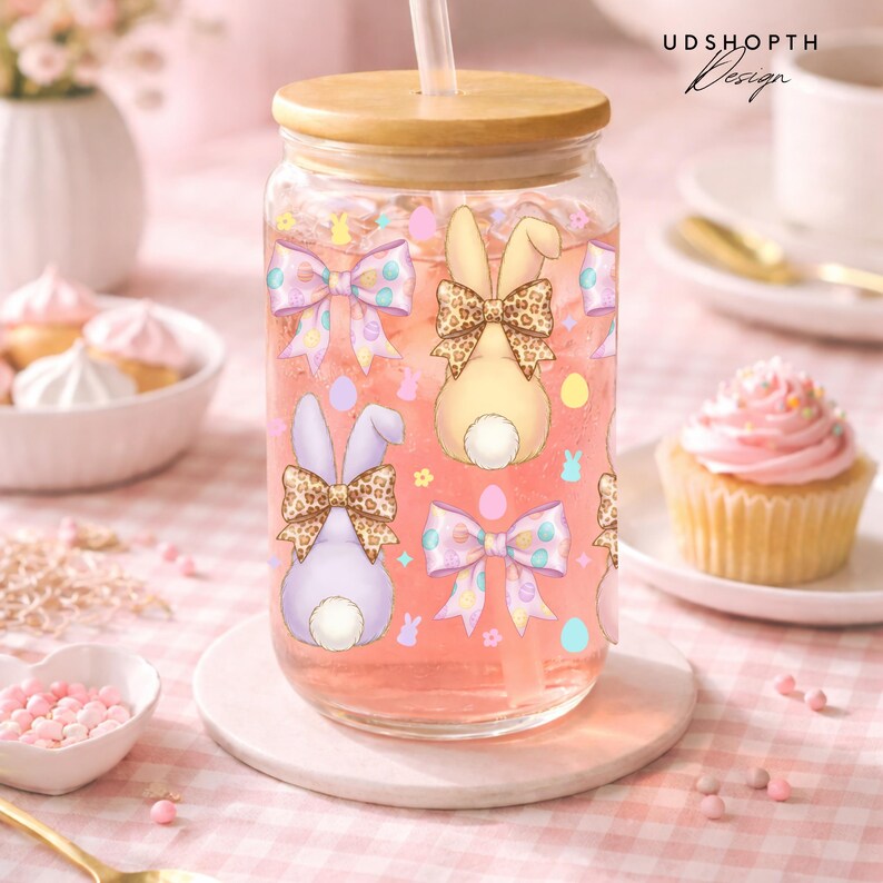 May include: A clear glass tumbler with a wooden lid and straw, filled with pink liquid. The glass is decorated with bunny and bow illustrations. The tumbler sits on a white coaster, with a pink and white checkered background.