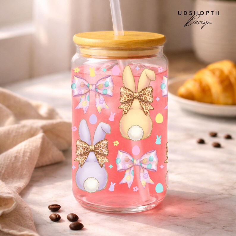 May include: A clear glass tumbler with a bamboo lid and straw, filled with pink liquid. The tumbler is decorated with Easter-themed bunny and bow illustrations. The design includes pastel colors and a playful aesthetic, perfect for spring.