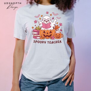 Pu&ograve; includere: Maglietta bianca con un fantasma dei cartoni animati che indossa gli occhiali e legge un libro. Il fantasma &egrave; seduto su una pila di libri e due zucche. Il testo "Spooky Teacher" &egrave; stampato sotto l'immagine.