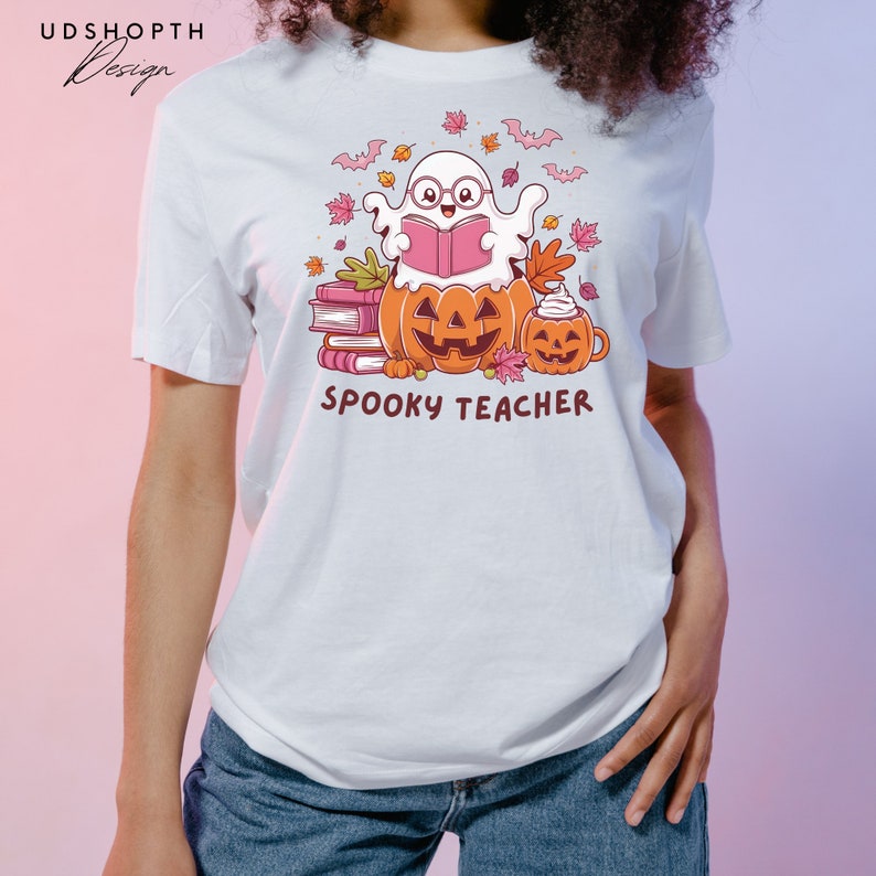 Pu&ograve; includere: Maglietta bianca con un fantasma dei cartoni animati che indossa gli occhiali e legge un libro. Il fantasma &egrave; seduto su una pila di libri e due zucche. Il testo "Spooky Teacher" &egrave; stampato sotto l'immagine.