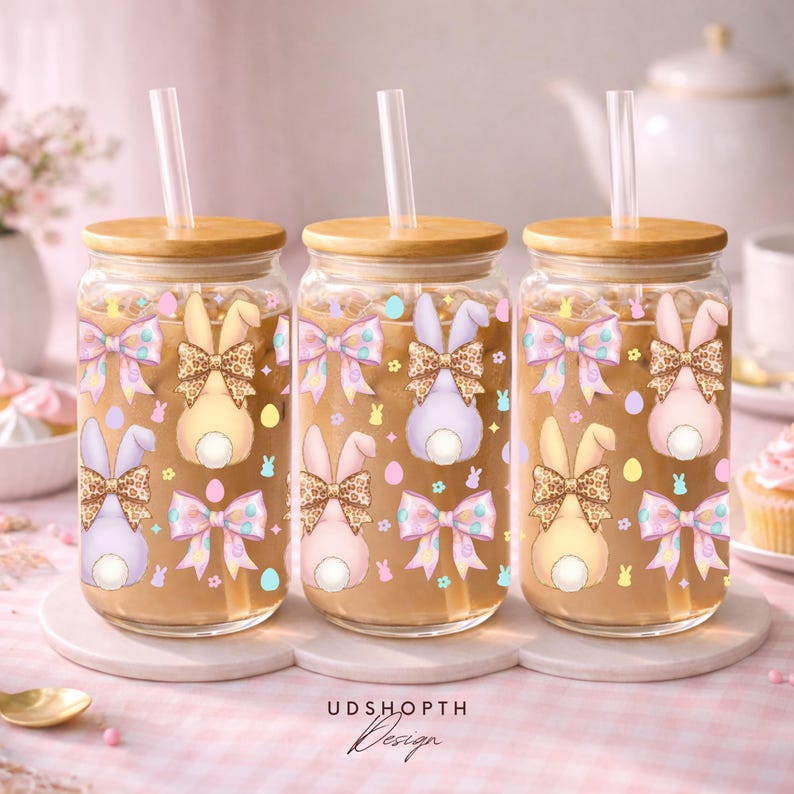 May include: Three glass tumblers with wooden lids and straws. Each tumbler features a bunny design with a bow and Easter egg accents. The tumblers are filled with a light brown liquid, perfect for cold beverages.
