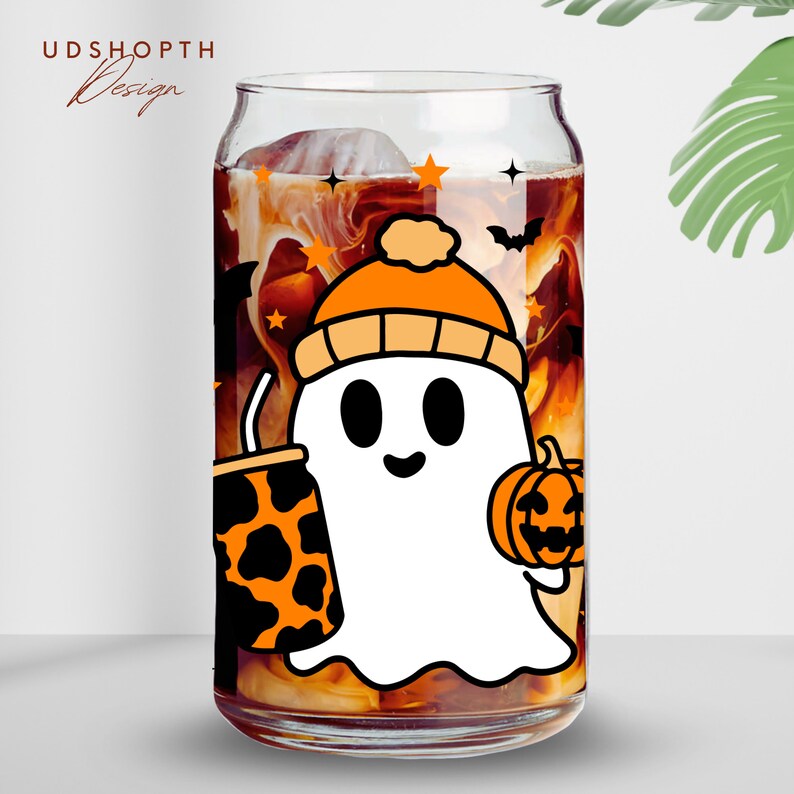 May include: A clear glass can with a white ghost wearing an orange and white hat holding a pumpkin and a black and orange patterned cup. The can is filled with a brown liquid and has a white and orange design with stars and bats.