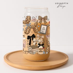 May include: A clear glass can-shaped tumbler with a light brown liquid inside, decorated with illustrations of books, bows, and the text "Coffee & Books." The design includes a balloon dog and the words "one more chapter."