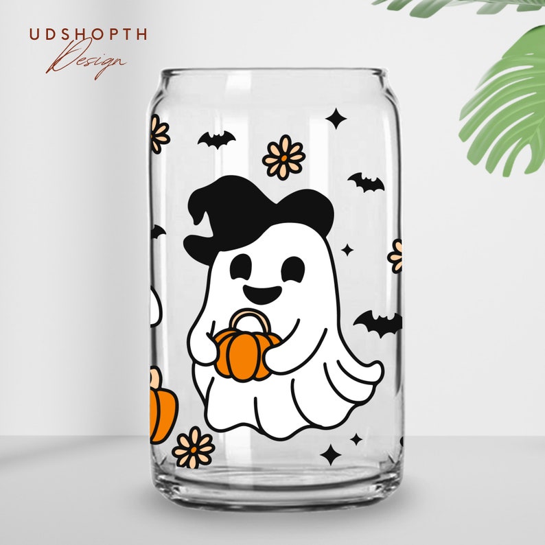 May include: A clear glass can with a white ghost wearing a black witch hat and holding an orange pumpkin. The ghost is surrounded by black bats, orange pumpkins, and yellow flowers.