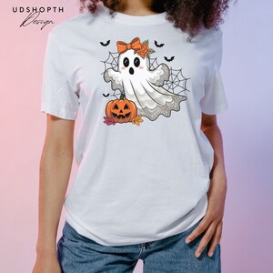 Pode incluir: Camiseta branca com um fantasma de desenho animado fofo usando um la&ccedil;o e segurando uma ab&oacute;bora de Halloween. O fantasma est&aacute; cercado por teias de aranha e morcegos.