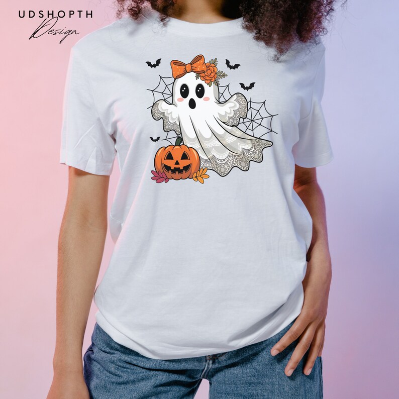 Pode incluir: Camiseta branca com um fantasma de desenho animado fofo usando um la&ccedil;o e segurando uma ab&oacute;bora de Halloween. O fantasma est&aacute; cercado por teias de aranha e morcegos.