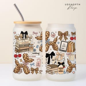 May include: Frosted glass can-shaped tumbler with a bamboo lid and a stainless steel straw. The tumbler features a whimsical design with books, bows, cherries, and a balloon dog. The words "READ MORE", "COZY CHAPTERS", and "COFFEE & BOOKS" are visible.