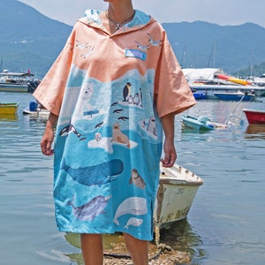 May include: A person wearing a hooded beach towel with a colorful ocean animal print. The towel is blue and orange with a variety of sea creatures, including whales, dolphins, penguins, and fish. The person is standing on a wooden dock with a body of water in the background.